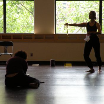 Joanna Garfinkel & Natalie Schneck in rehearsal. One standing with their hand out, the other sitting on the floor