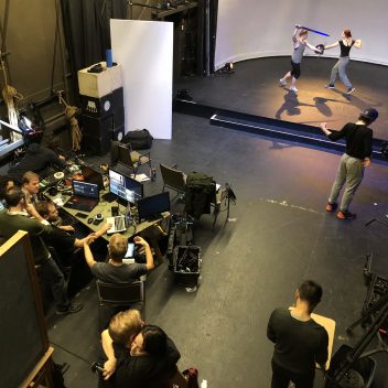 A rehearsal hall shot from above. There are two people sword fighting, and many others watching around filming equipment