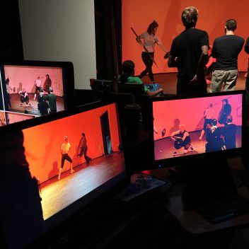 A small group gathered, rehearsing with a red background. There are a few monitors, which are showing various angles of those rehearsing.