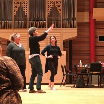 Three people in rehearsal, one is pointing up at something. The other two are looking at that person. The walls are wooden and there is a table and some chairs