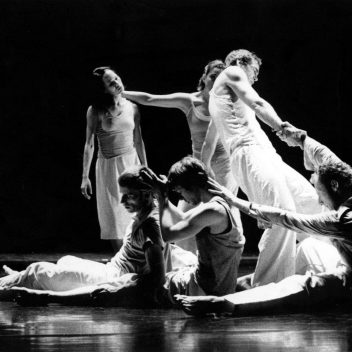 A group of people in movement. Some are seated on the floor, others are standing. They are all connected, by their arms. It’s in black and white.