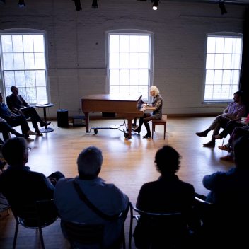 Jane Miller seated, playing a piano. A circle of people are seated around the piano, listening. The circle of people are in darkness and Jane is lit up.