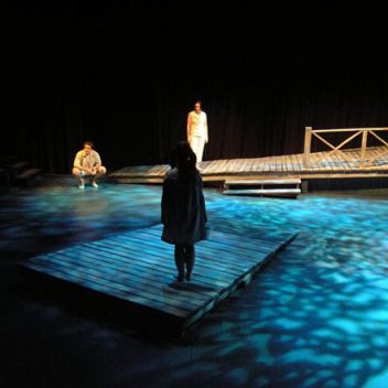 Three people on stage, with blue light on the floor. One is sitting on their heels, facing the front. One is standing on a wooden dock facing forward. The other is standing on a different wooden dock, with their back to the front.