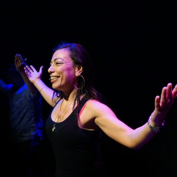 Carmen Aguirre standing with their hands outstretched, behind is a black background