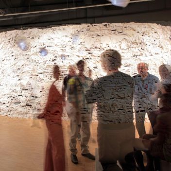 A group of people in a circle, some are seated but the rest are standing. Their bodies are translucent, so a textured wall can be seem