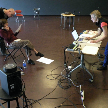 Three people in a rehearsal hall. One is sitting looking at a paper they are holding, one is behind them, also looking at that paper. One person is standing behind a table, looking at a laptop